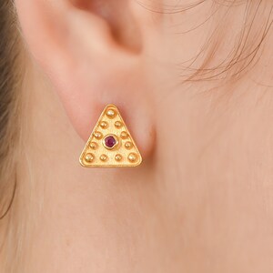 May include: Gold triangle earrings with a small red gemstone in the center. The earrings have a textured surface.