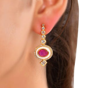 May include: A gold-tone drop earring featuring a vertical row of four small clear stones leading to a central oval-shaped pink stone surrounded by a white enamel border and a halo of tiny clear stones, finished with a single square clear stone at the base.