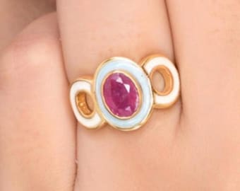 Puede incluir: Un anillo de oro con una piedra preciosa ovalada de color rosa intenso en el centro, rodeada por un borde de esmalte azul claro. El diseño incluye dos bucles circulares de oro abiertos a los lados, con detalles en esmalte blanco.