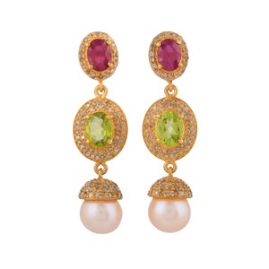 May include: Gold-tone drop earrings featuring a ruby, peridot, and pearl design. The earrings have an oval ruby at the top, a peridot in the middle, and a pearl at the bottom. The gemstones are surrounded by small, sparkling accents.