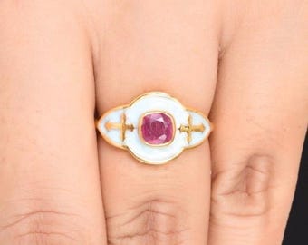 Puede incluir: Un anillo de oro con una piedra preciosa de color rojo rosado en talla cojín, engastada en una placa ovalada de esmalte blanco. La superficie esmaltada está decorada con dos cruces doradas a los lados, enmarcada por un borde de oro festoneado.