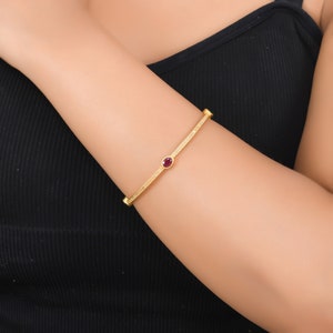 Peut inclure: Un bracelet jonc doré avec une surface texturée et une pierre précieuse rouge foncé ovale au centre. Le bracelet est porté sur un bras, et l'arrière-plan est un débardeur noir.