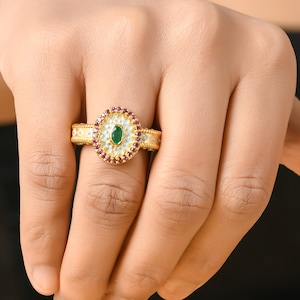 May include: A gold ring featuring an oval, emerald-colored gemstone at the center, surrounded by small red gemstones and a light blue enamel design. The ring's band has a detailed pattern with white and gold accents.