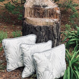 Pu&ograve; includere: Tre cuscini da lancio grigi e bianchi con un design a foglie sottile sono disposti su un ceppo d'albero in un ambiente naturale all'aperto.