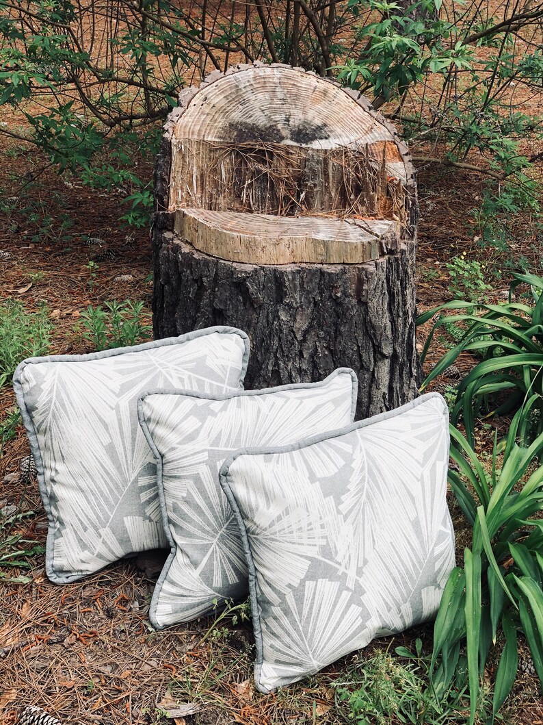 Pu&ograve; includere: Tre cuscini da lancio grigi e bianchi con un design a foglie sottile sono disposti su un ceppo d'albero in un ambiente naturale all'aperto.