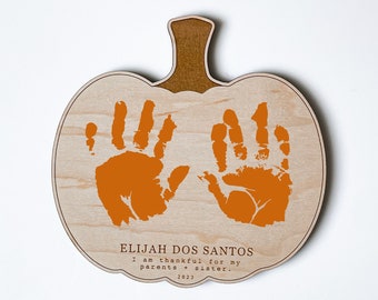 May include: A wooden pumpkin-shaped ornament with two orange handprints and the text "ELIJAH DOS SANTOS I am thankful for my parents + sister. 2023."