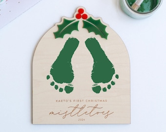 May include: A wooden sign with a green holly leaf design and two green footprints. The sign reads "Kaeto's First Christmas Mistletoes 2024".