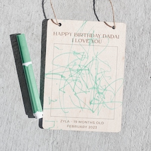 May include: A wooden sign with the text "Happy Birthday Dadai I Love You" and a green drawing inside a frame. The sign also includes the text "Zyla - 19 months old February 2023".