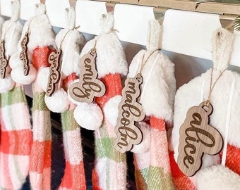 May include: A set of eight personalized stockings with names written on wooden tags. The stockings are white with red and green plaid patterns and are hung on a white mantel.