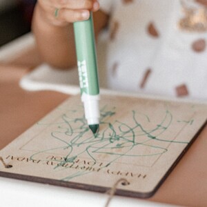 May include: A child's hand holding a green marker, drawing on a wooden board with the words "Have a wonderful day!" written on it.