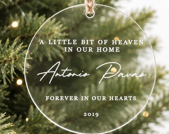 May include: Clear, round Christmas ornament with the text "A LITTLE BIT OF HEAVEN IN OUR HOME" and "Antonio Pavas" in white script. The ornament also includes "FOREVER IN OUR HEARTS" and the year "2019". A brown twine loop is at the top.
