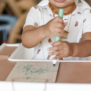 May include: A young child sits at a table, drawing with a green marker on a piece of paper. The child is wearing a white shirt with brown spots and a gold necklace with an amber pendant.