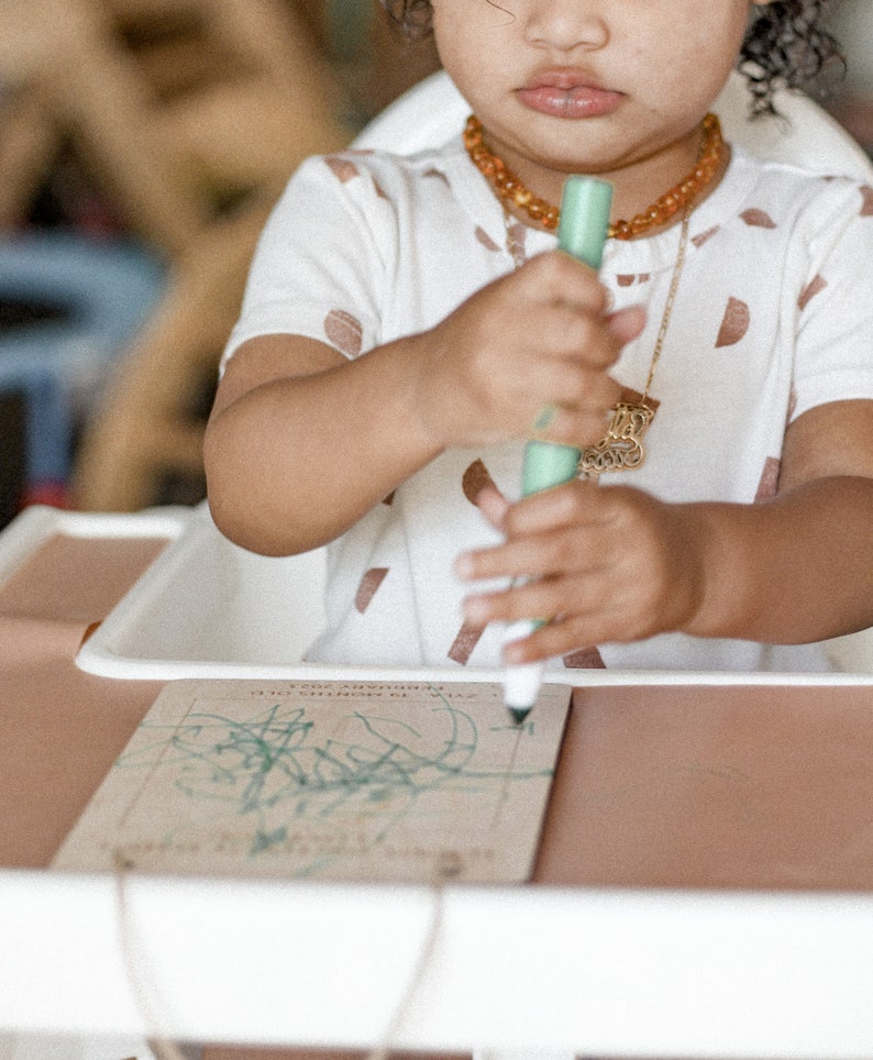 May include: A young child sits at a table, drawing with a green marker on a piece of paper. The child is wearing a white shirt with brown spots and a gold necklace with an amber pendant.