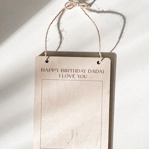 May include: A brown wooden sign with a white border and twine hanging from the top. The sign says "Happy Birthday Dadai I Love You" and "Zyla - 19 Months Old February 2023".
