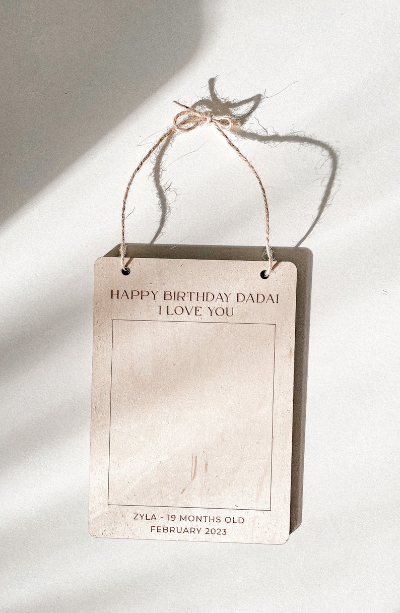 May include: A brown wooden sign with a white border and twine hanging from the top. The sign says "Happy Birthday Dadai I Love You" and "Zyla - 19 Months Old February 2023".