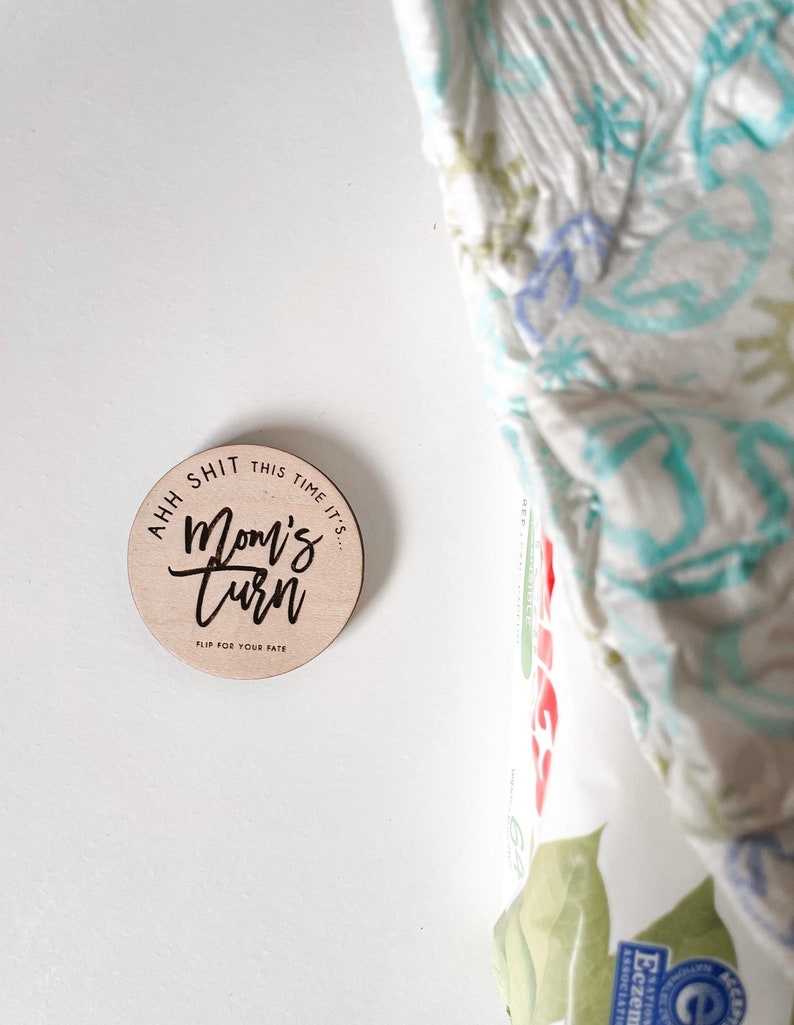 May include: A round wooden disc with the text "Ahh shit this time it's... Mom's Turn. Flip for your fate" in black script. The disc is light brown and sits on a white surface. A diaper package is partially visible in the background.