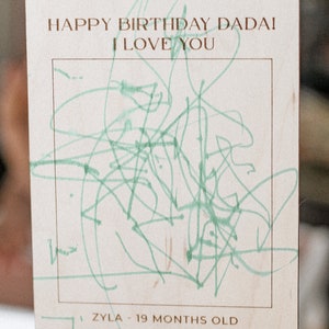 May include: A wooden sign with the text "Happy Birthday Dada! I Love You" and a green drawing inside a frame. The sign also includes the text "Zyla - 19 months old February 2023".