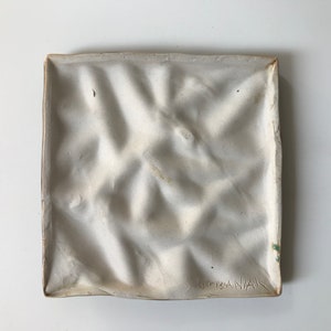 May include: White ceramic tile with a textured surface and a raised pattern. The tile is signed "GRISANAK" in the bottom right corner.