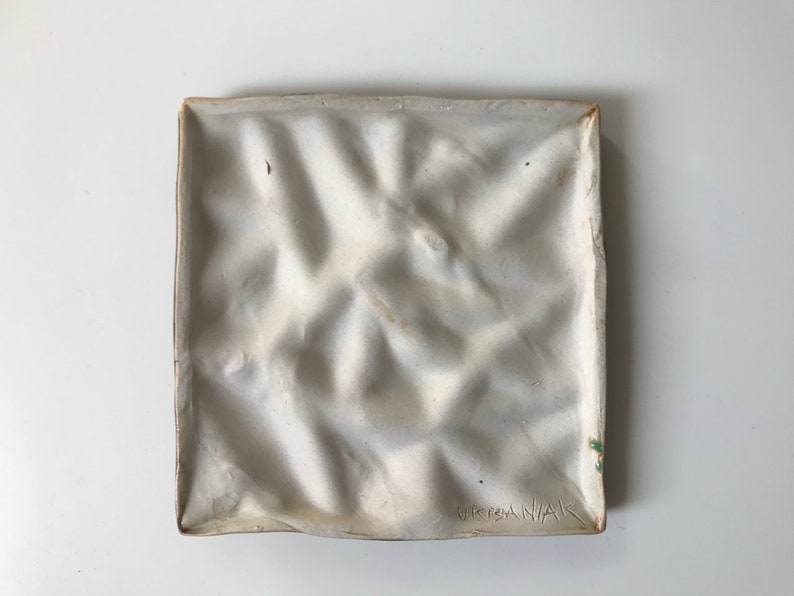 May include: White ceramic tile with a textured surface and a raised pattern. The tile is signed "GRISANAK" in the bottom right corner.