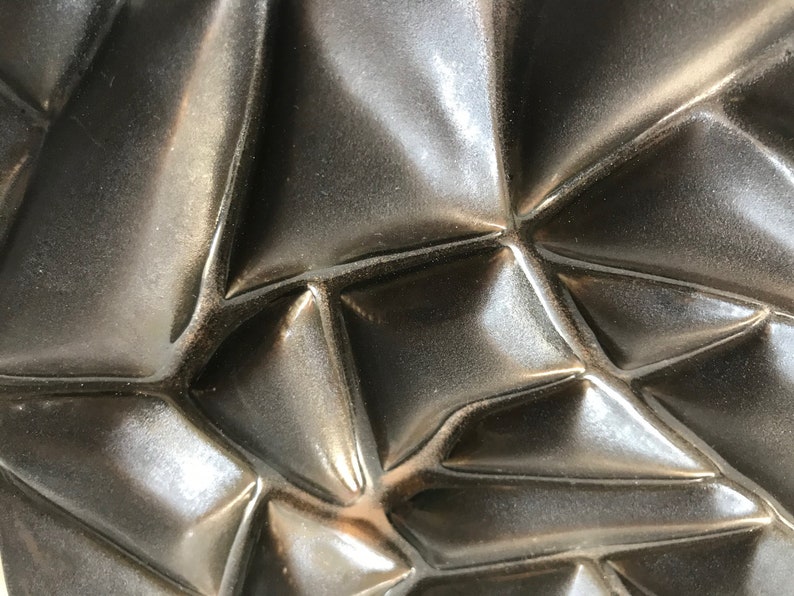 May include: Close-up of a dark gray, textured surface with a geometric pattern. The surface is made of a material that appears to be ceramic or metal.