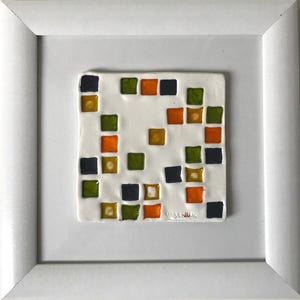 Op de afbeelding: Een vierkante witte keramische tegel met een verspreid patroon van kleine, glanzende, verhoogde vierkantjes in tinten donkerblauw, olijfgroen en oranje, geplaatst in een eenvoudige witte lijst. De naam "URBANIAK" staat rechtsonder in reliëf.