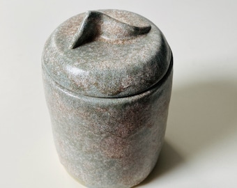 May include: A ceramic jar with a lid featuring a unique, abstract design. The jar is a mottled green and brown color with a speckled finish.
