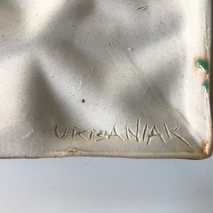 May include: Close-up of a white ceramic plate with a textured surface. The plate has a green glaze on the edge and the signature "Urbaniak" is inscribed on the bottom.