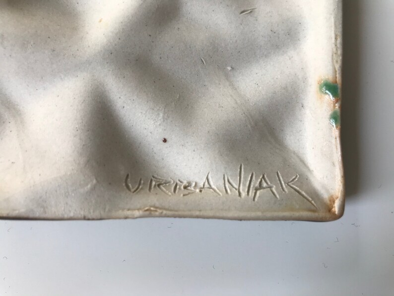 May include: Close-up of a white ceramic plate with a textured surface. The plate has a green glaze on the edge and the signature "Urbaniak" is inscribed on the bottom.