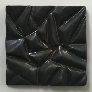 May include: A black ceramic tile with a textured, geometric pattern. The tile has a glossy finish and a raised, three-dimensional design.