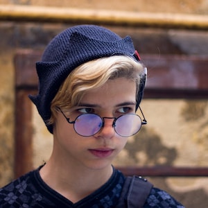 Può includere: Un giovane che indossa un berretto nero con un accento rosso e occhiali rotondi colorati di blu. Indossa una camicia a motivi blu scuro e neri.
