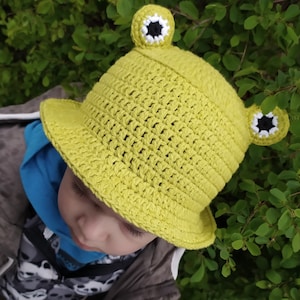 Op de afbeelding: Een limoengroene gehaakte bucket hat met twee uitstekende kikkerogen aan de bovenkant. De ogen zijn gemaakt met witte en zwarte garen details. De hoed heeft een gestructureerd weefpatroon en een brede, naar beneden hellende rand rondom.