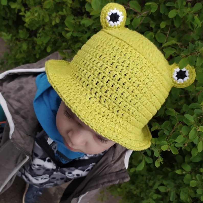 Op de afbeelding: Een limoengroene gehaakte bucket hat met twee uitstekende kikkerogen aan de bovenkant. De ogen zijn gemaakt met witte en zwarte garen details. De hoed heeft een gestructureerd weefpatroon en een brede, naar beneden hellende rand rondom.