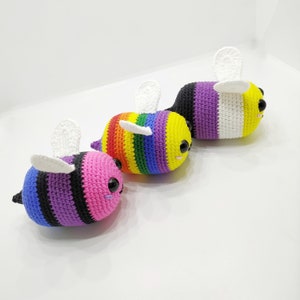 May include: Three crocheted bee plushies in a row. The bee on the left is black, purple, and white. The middle bee is rainbow coloured. The bee on the right is black, purple, and white.