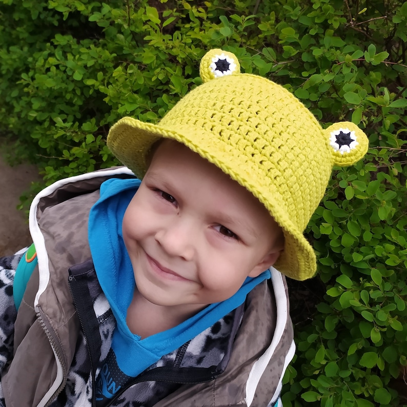 Op de afbeelding: Een limoengroene gehaakte bucket hat met twee uitstekende kikkerogen aan de bovenkant. De ogen zijn gemaakt met wit, zwart en limoengroen garen, wat een cirkelvormig patroon cre&euml;ert dat het uiterlijk van kikkerogen op de kroon van de hoed nabootst.