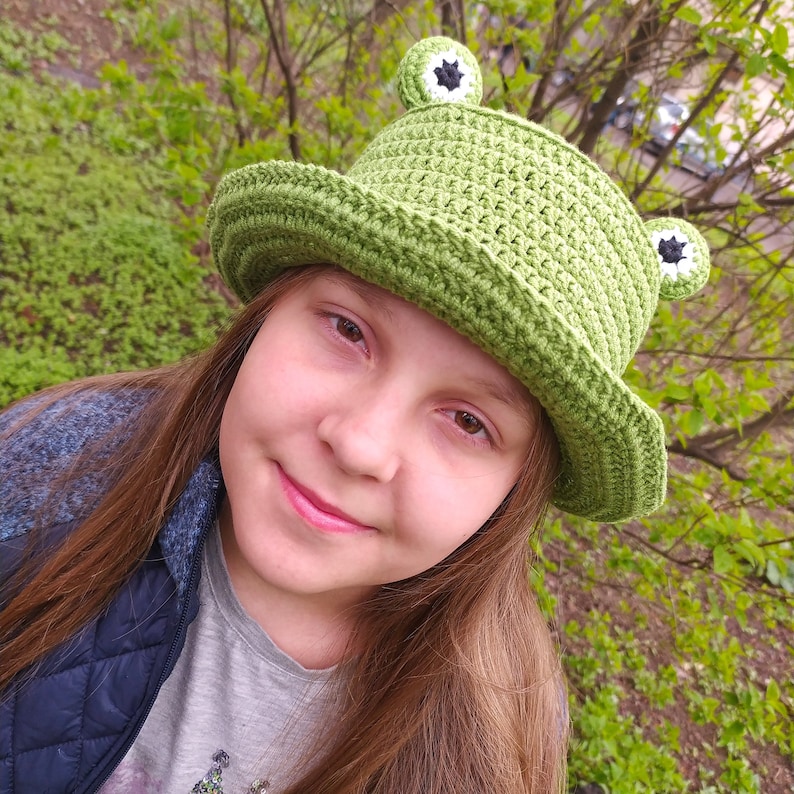 Op de afbeelding: Een groene gehaakte bucket hat met twee uitstekende kikkerogen aan de bovenkant. De ogen zijn cirkelvormig met witte randen, zwarte centra en groene buitenkanten. De hoed heeft een brede, naar beneden lopende rand en een textuur.