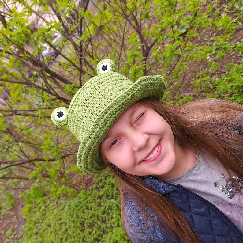Op de afbeelding: Een felgroene gehaakte bucket hat met twee uitstekende kikkerogen aan de bovenkant. De ogen zijn cirkelvormig met wit garen, zwarte centra en groene randen. De hoed heeft een gestructureerd ribbelpatroon en een brede, neerwaartse rand.