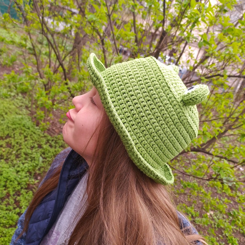 Op de afbeelding: Een felgroene gehaakte bucket hat met twee kleine, uitstekende kikkerogen aan de bovenkant. De hoed is gemaakt met een dicht, gestructureerd steekpatroon en heeft een brede, licht neerwaartse rand die de gehele kroon omcirkelt.