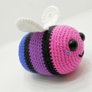 Op de afbeelding: Een gehaakt bij-speelgoed met een gestreept lichaam in roze, paars, zwart en blauw. De bij heeft witte vleugels en zwarte ogen.