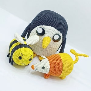 Pu&ograve; includere: Tre peluche realizzati a mano all'uncinetto: un pinguino grigio scuro e bianco con il becco giallo, un'ape a strisce gialle e nere con ali bianche e una piccola creatura simile a un topo con corpo bianco, arancione e giallo e coda.