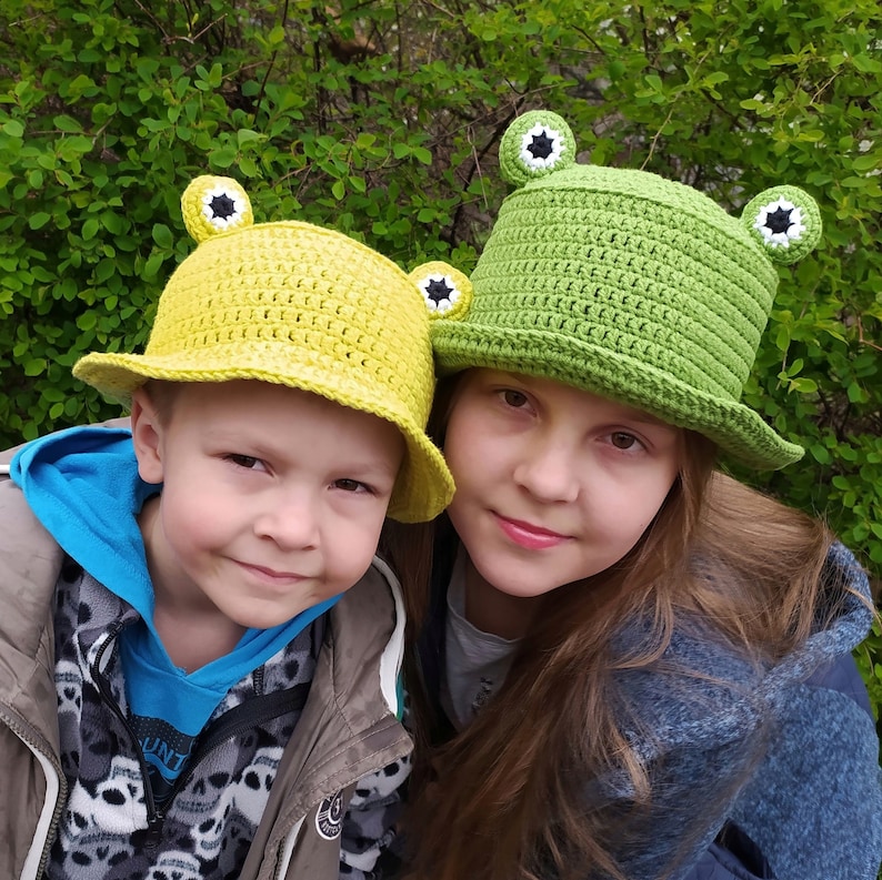 Op de afbeelding: Twee gehaakte bucket hats met kikkerontwerpen. De ene hoed is felgeel en de andere is grasgroen. Beide hoeden hebben een platte bovenkant, een brede rand en twee kleine ronde kikkerogen die aan de bovenkant zijn bevestigd met witte, zwarte en groene details.