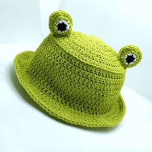 Op de afbeelding: Een limoengroene gehaakte bucket hat met twee uitstekende kikkerogen aan de bovenkant. De ogen zijn gemaakt met wit en zwart garen om pupillen na te bootsen. De hoed heeft een getextureerd uiterlijk met een brede, ronde rand.