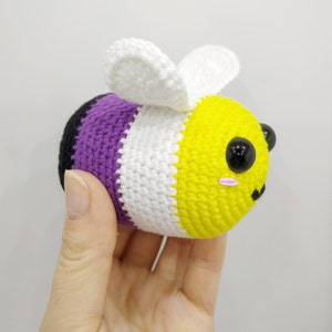 May include: Crocheted bee plush toy with purple, white and yellow stripes. The bee has black eyes and pink cheeks.