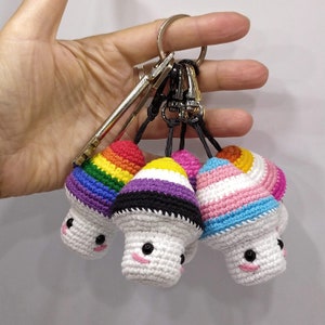 Może przedstawiać: Zestaw pięciu breloków w kształcie grzybów wykonanych na szydełku, każdy z innym wzorem kolorystycznym reprezentującym flagę dumy. Breloki są przymocowane do srebrnych kółek do kluczy.
