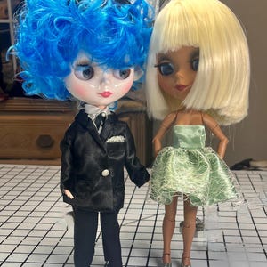 May include: Two fashion dolls. One doll has blue curly hair, wears a black suit, and has a white shirt and tie. The other doll has blonde hair, a green dress, and silver shoes. They stand on a black and white checkered surface.