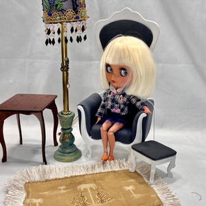 Puede incluir: Una escena de casa de muñecas con una muñeca sentada en un sillón en miniatura, una lámpara de pie con una pantalla decorativa, una mesa pequeña, una alfombra con flecos y un taburete. La muñeca lleva una blusa a cuadros y una falda azul.