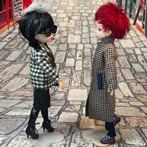 May include: Two fashion dolls are posed on a cobblestone street. One doll wears a black and white checkered jacket, black skirt, and heels. The other doll wears a long checkered coat and blue shoes. Both dolls have unique hairstyles and accessories.