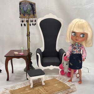 Może przedstawiać: Miniaturowa scena wnętrza z krzesłem z wysokim oparciem, małym stolikiem, lampą, dywanem i lalką. Krzesło jest czarno-białe, stół jest brązowy, a lampa ma dekoracyjny klosz. Lalka ma na sobie kwiatowy top i ciemną spódnicę.
