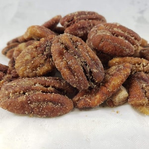 May include: A close-up shot of a pile of candied pecans. The pecans are a rich brown color, coated in a light-colored, granular sugar. The nuts are arranged in a slightly overlapping heap, suggesting a snack or treat.