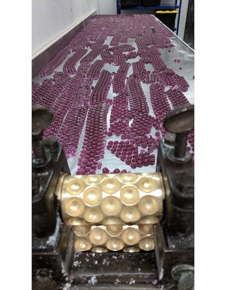May include: A close-up of a candy making machine with a gold roller and a large amount of pink candy on a metal surface.