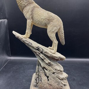 May include: A detailed sculpture of a howling wolf, standing on a rocky outcropping. The wolf is a light beige color, with textured fur. The base of the sculpture is a square shape. The text "JACK KOWEN" is visible on the base.